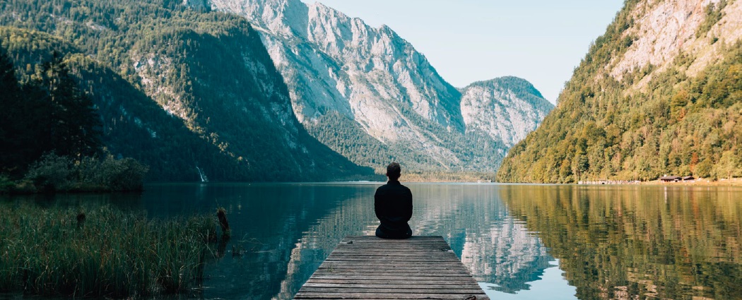 Someone sitting on a dock in-front of a mountain lake.