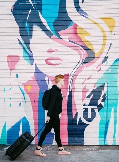 Someone with a suitcase in-front of a graffiti'd wall.