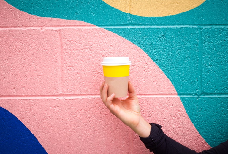 A yellow coffe cup held in-front of a colourful mural.